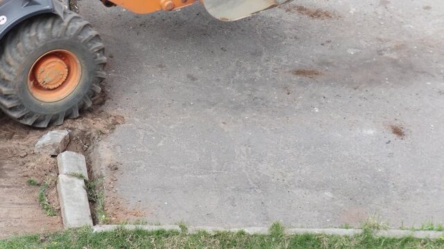 Orange heavy construction machinery at work, front loader bucket and large wheels on asphalt road and dirt, clearing earth near concrete curb, outdoor daytime activity.