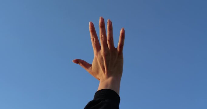 Raised hand waves against clear blue sky. Person reaches upward in bright daylight. Gesture suggests greeting, freedom, hope, or asking for help. Minimal outdoor footage offers useful copy space.