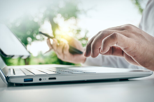 Close up of businessman hands using smartphone and typing on laptop computer in outdoor green nature setting, concept of remote work, mobile technology, and freelance productivity.