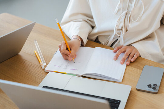A young woman sits at a wooden desk and writes notes in a lined notebook with a yellow pencil. Laptops and a smartphone rest nearby while she focuses on her daily tasks.