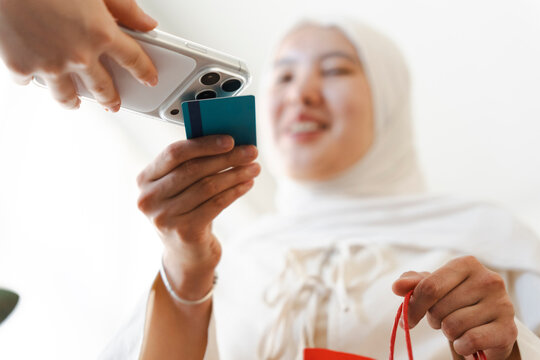 A smiling woman wearing a hijab pays for her purchases using a mobile phone and credit card. She holds a shopping bag while completing a quick contactless transaction.