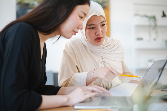 Two young women work together in a bright office. One woman with long dark hair types on a laptop while her colleague wearing a hijab points at the screen with a pencil.