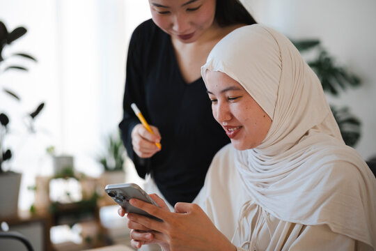 Two diverse women collaborate in a bright office. A female worker wearing a hijab uses a smartphone while her colleague stands beside her, looking at the screen and smiling.