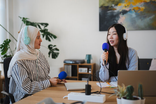 Two young women are recording an audio podcast in a bright home studio. They wear headphones and hold microphones, smiling while discussing a topic across a wooden desk.