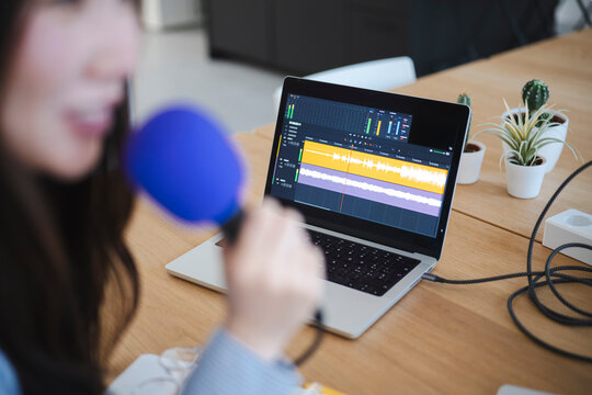 A young woman speaks into a blue microphone while recording a podcast in a bright office. A laptop on the wooden desk displays an active audio waveform editing software.
