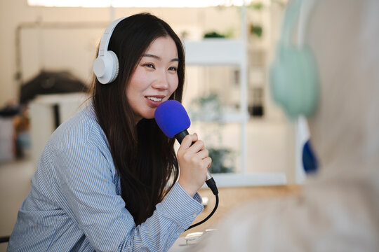 A young Asian woman wearing white headphones speaks into a blue microphone while recording an audio podcast interview in a brightly lit modern office workspace.