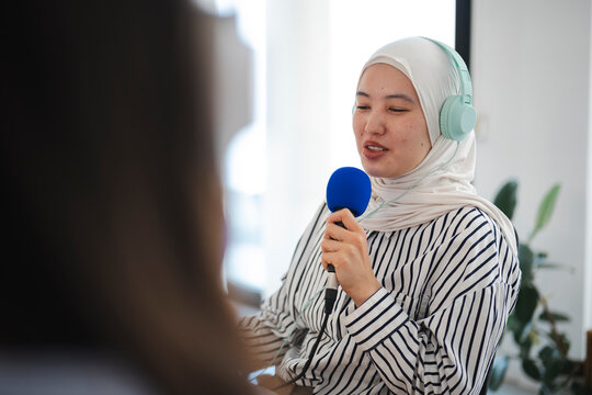 A young woman wearing a hijab and headphones speaks into a blue microphone during an interview in a bright office. Her colleague listens attentively off-camera.
