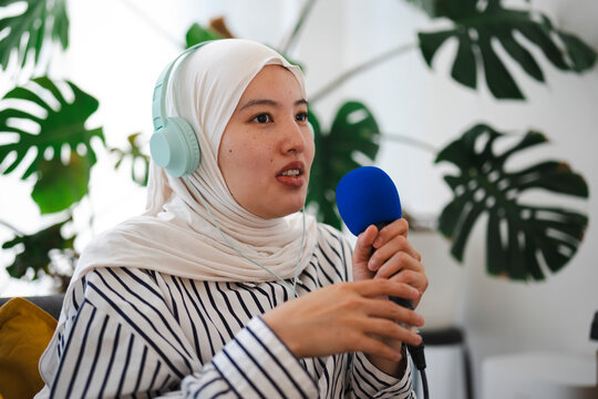 A young woman wearing a white hijab and headphones speaks directly into a blue microphone while recording a podcast in a bright room filled with green plants.