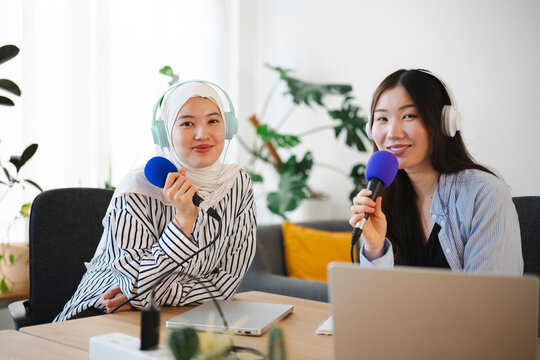 Two young female podcasters wearing headphones sit at a wooden table in a bright room. They hold microphones and smile at the camera while recording their audio show.