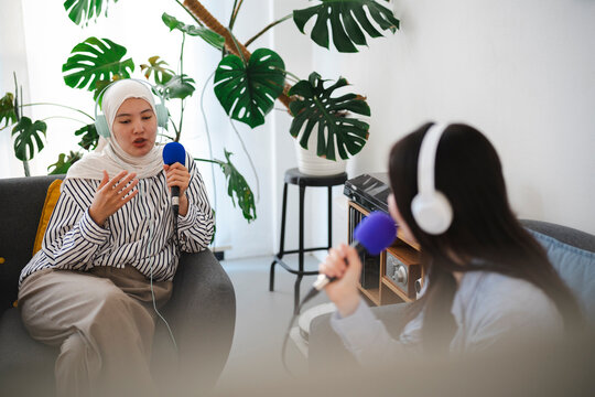 Two young women are sitting on a couch in a bright room, wearing headphones and holding microphones while recording an interview for their online podcast.