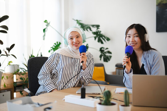 Two diverse young women wearing headphones and holding microphones while recording a podcast together in a bright modern studio. They smile at the camera.
