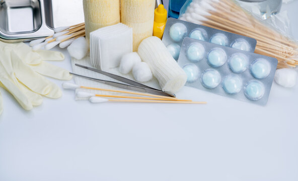 Wound care supplies with gauze rolls, cotton swabs, gloves and metal forceps on a white surface, alongside dressing pads, packaged cotton balls, small bottles and a divided medical tray.