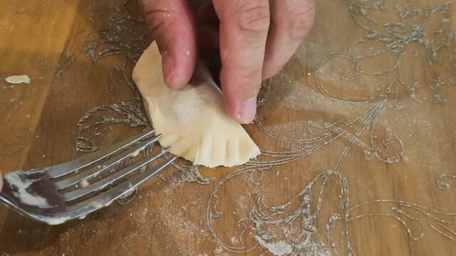 Sealing homemade dumpling edges with metal fork creating decorative crimped pattern on dough. Metal fork seals raw dumpling dough edge creating neat decorative crimped pattern on table.