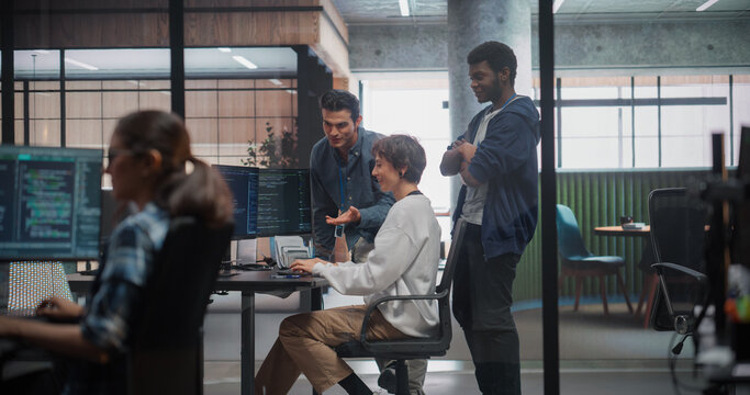 Multiethnic Software Development Team Meeting and Talking Near a Desk With Code on Screens in a Spacious Tech Office, Female Coder Shares Ideas While Two Male Engineers Listen, Using Computer