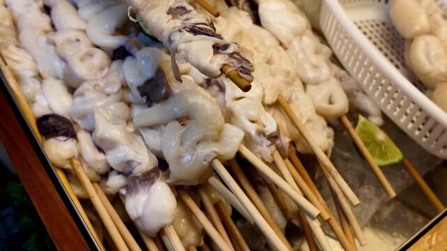 Close up shot of fresh raw squid skewers arranged on a display counter at a traditional street food market stall in Thailand.
