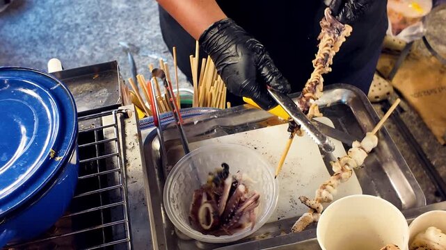 Close up shot of street food vendor hands in black gloves using tongs to remove freshly grilled squid pieces from a wooden skewer into a plastic bowl.
