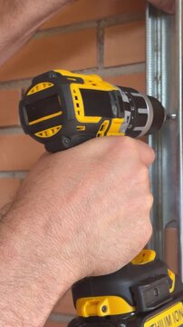 A person operates a power drill to secure a metal frame to the wall in a workshop setting. The task focuses on drilling and fastening.