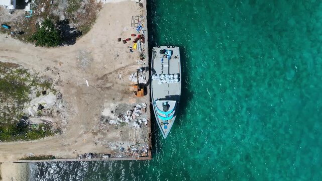High angle top-down drone perspective of boat moored at island harbour dock bordering clear blue ocean. Ideal visual asset for marine logistics and travel content.