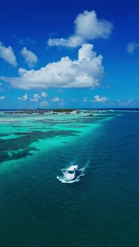 Drone shot of white boat leaving water wake in deep blue sea near tropical paradise island with resort. Perfect vertical asset for travel reels and promo videos.Maldives.Vertical video