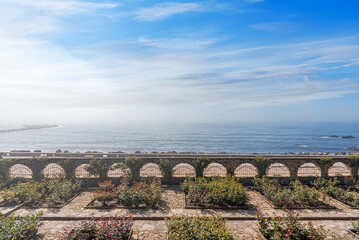 Naklejka premium A beautiful seaside garden features manicured flower beds and an arched stone wall overlooking a calm blue sea under a bright, misty sky.