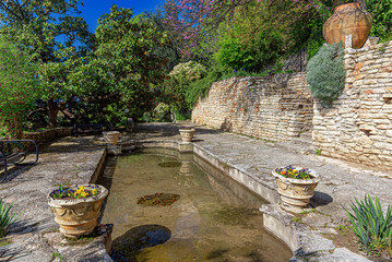 Naklejka premium A serene stone courtyard features a reflective decorative pond surrounded by potted flowers and ancient stone walls under a clear blue sky.