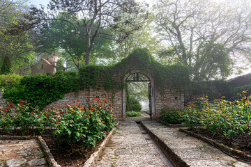 Naklejka premium A stone archway covered in green vines stands in a peaceful garden with blooming rose bushes and lush trees.
