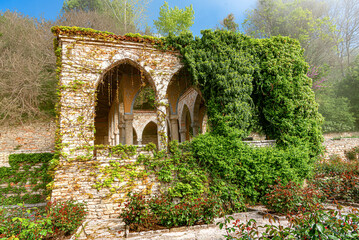 Naklejka premium An ancient stone pavilion with arched openings covered in lush green ivy stands in a sunny garden.