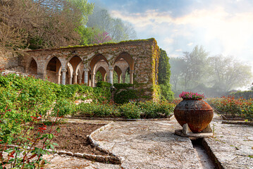 Naklejka premium A beautiful stone archway covered in ivy overlooks a lush green garden with a large decorative vase filled with pink flowers under a bright sky.