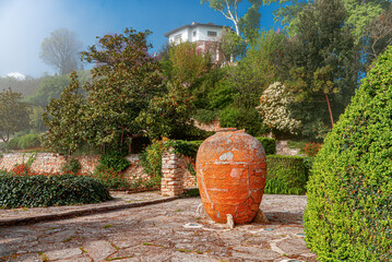 Naklejka premium A large clay amphora stands in a scenic Mediterranean garden with stone walls, lush green foliage, and a white villa on a misty hill.