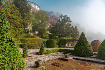 Naklejka premium A beautiful manicured garden features green topiary bushes, stone pathways, and a foggy background on a sunny morning.
