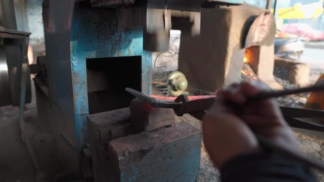 Close-up of a blacksmith using tongs to hold heated metal on an anvil while a mechanical power hammer strikes it in a traditional metalworking workshop.