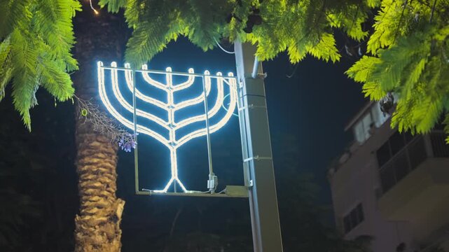 Festive Hanukkah menorah street decoration brightly illuminating a city street at night, with palm tree fronds and foliage framing the traditional Jewish symbol of the holiday