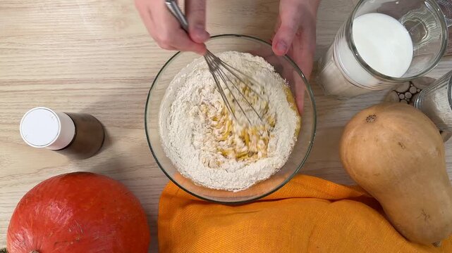 Hands are working with ingredients in a clear glass bowl containing a yellow mixture alongside dry goods, the setup sits on a wooden surface near pumpkins and milk bottles