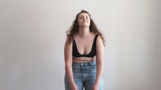 Body insecurity and self doubt, young curvy woman in black bra and jeans touching belly and sighing on white background. Body image struggle concept, woman looks up, down and at her stomach