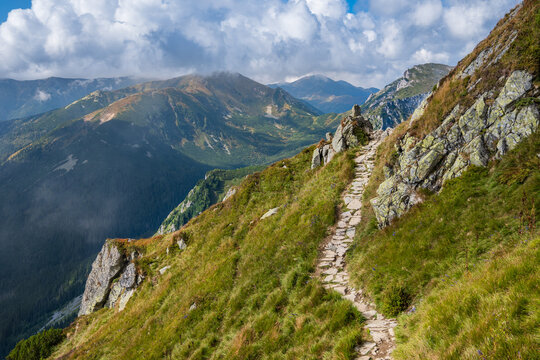 A precarious stone path winds along a narrow, rugged mountain ridge, offering dramatic views of deep valleys and hazy distant mountains in the Tatras Mountains in Zakopane, Poland.