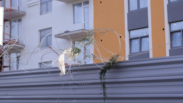Spirals of tangled barbed wire rest atop a corrugated metal barrier in front of a modern apartment building with white and orange facades