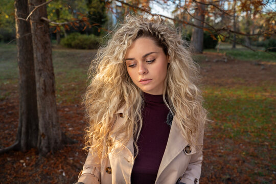 A young woman with curly blonde hair looks down with a calm expression while standing in an autumn park.