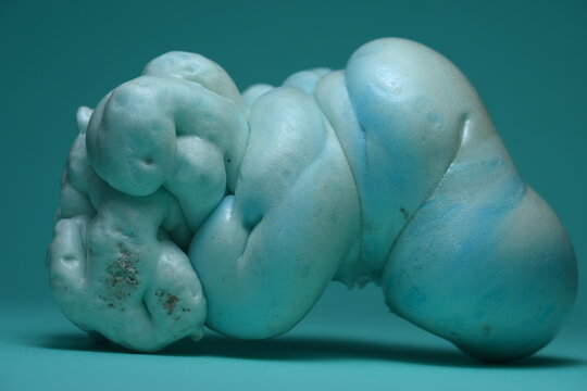A studio close-up of an abstract biomorphic sculpture made of expanded foam, showing light blue and turquoise organic shapes against a solid teal background.