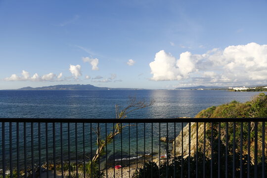 A scenic view of the blue ocean and distant coastline seen through a balcony railing in Okinawa.