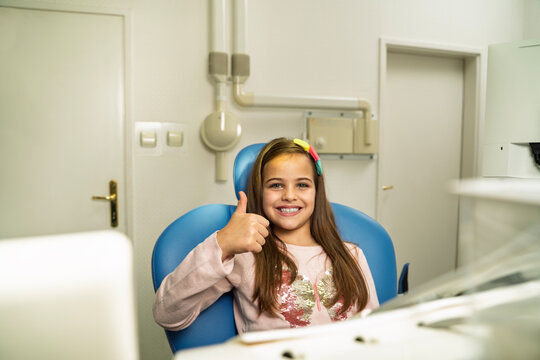 A smiling young girl gives a thumbs-up while sitting in a blue dental chair inside a clinic