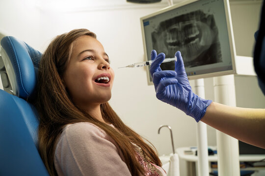 A young girl smiles in a dental chair as a dentist prepares an anesthesia injection in front of a digital dental X-ray monitor