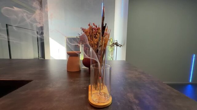 Thin white smoke rising from burning incense sticks in glass holder on dark table. Cozy modern interior background with dried flowers and neon light. Aroma therapy, spa, zen, meditation, relaxation.
