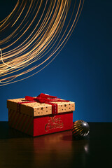 Fototapeta premium Christmas gift box with red ribbon and holiday bauble on dark background with golden light trails