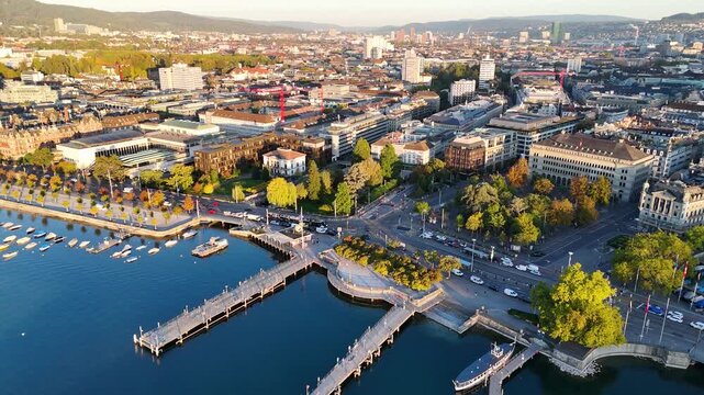 Drone glides over deep blue Lake Zurich toward Zurich. View of lakeside houses, sailboats, and distant Zurich skyline on a bright, calm day.