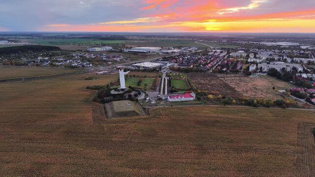 Aerial drone footage of the monumental Christ the King statue in Swiebodzin, Poland, surrounded by open fields and urban development beneath a colorful sunset sky. A wide cinematic view