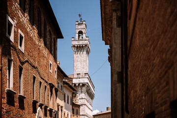 Naklejka premium Historic town hall tower of Palazzo Comunale in Montepulciano, Tuscany, Italy, viewed through a narrow street lined with old brick buildings in the medieval town center.