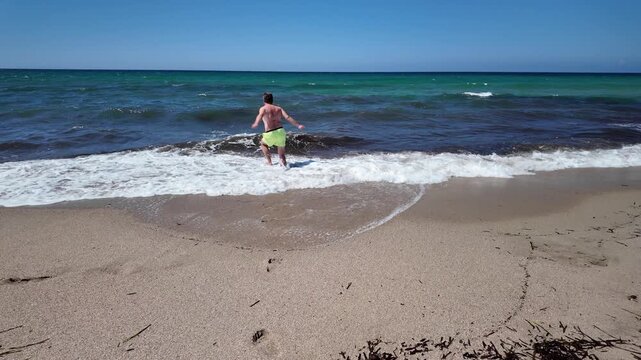 A middle-aged man in bright swimming trunks jumping into the turquoise sea at a sandy beach.