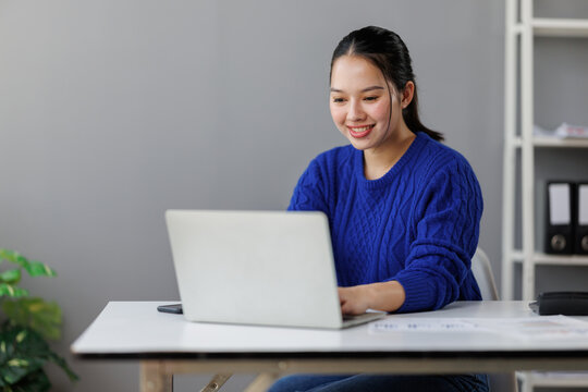 Asian woman working remotely on laptop, smiling and engaging in online business communication, showcasing freelancer flexibility and digital entrepreneurship in a modern office