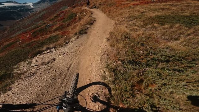 First-person POV footage of a mountain biker descending a rugged alpine trail with another cyclist riding ahead, surrounded by steep mountain terrain and dramatic scenery.