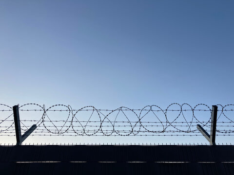 Silhouette of razor wire and barbed wire on a security fence against a clear blue sky